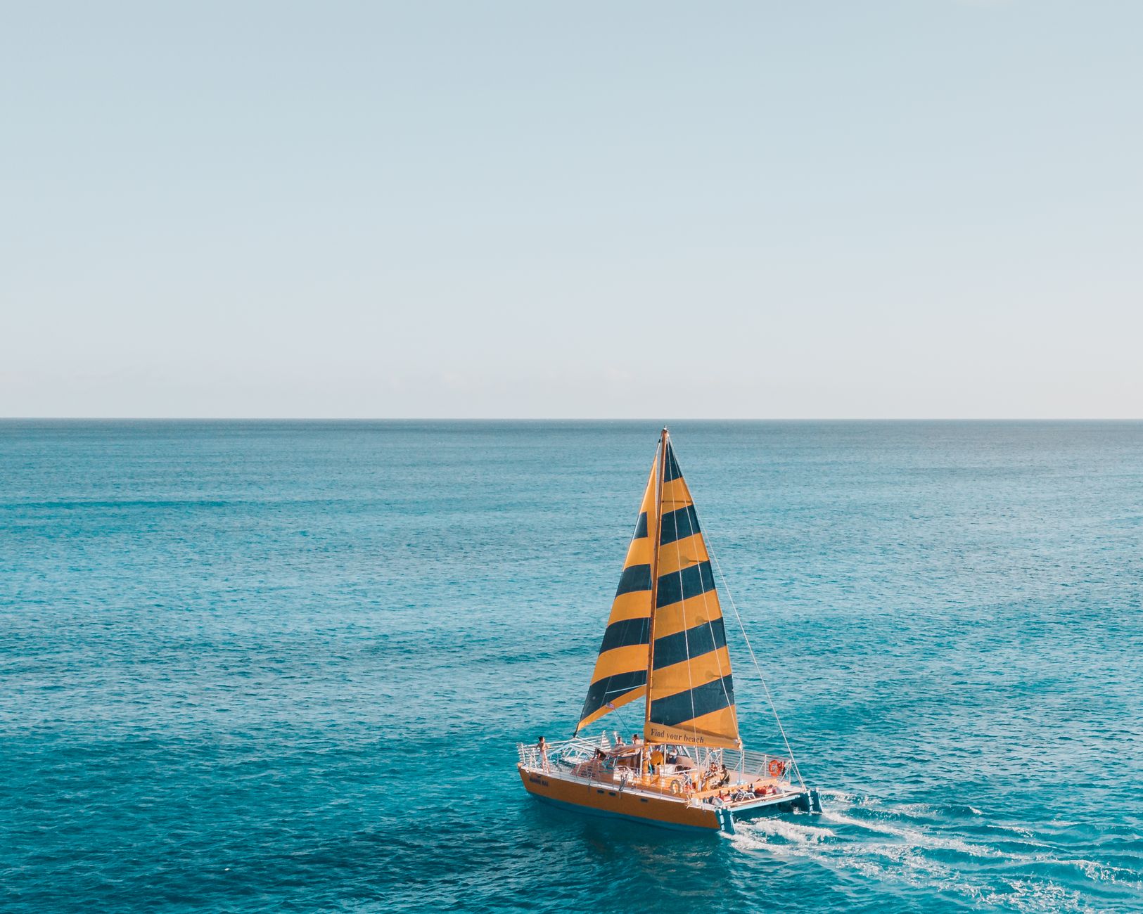 A striking orange sailboat with striped sails navigates the peaceful blue ocean during daylight.