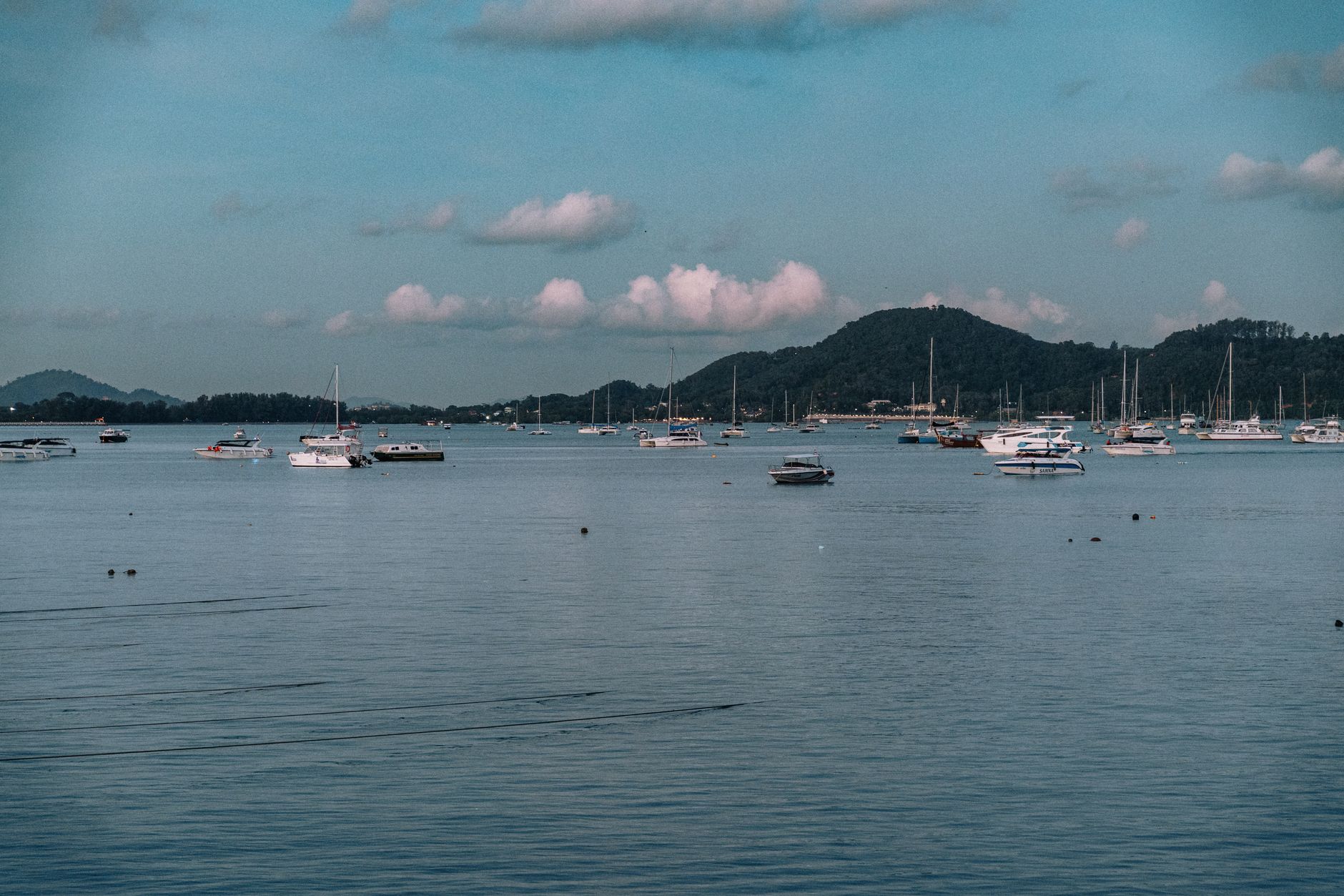 Yachts float peacefully on a calm bay against a hilly backdrop in Phuket, Thailand.